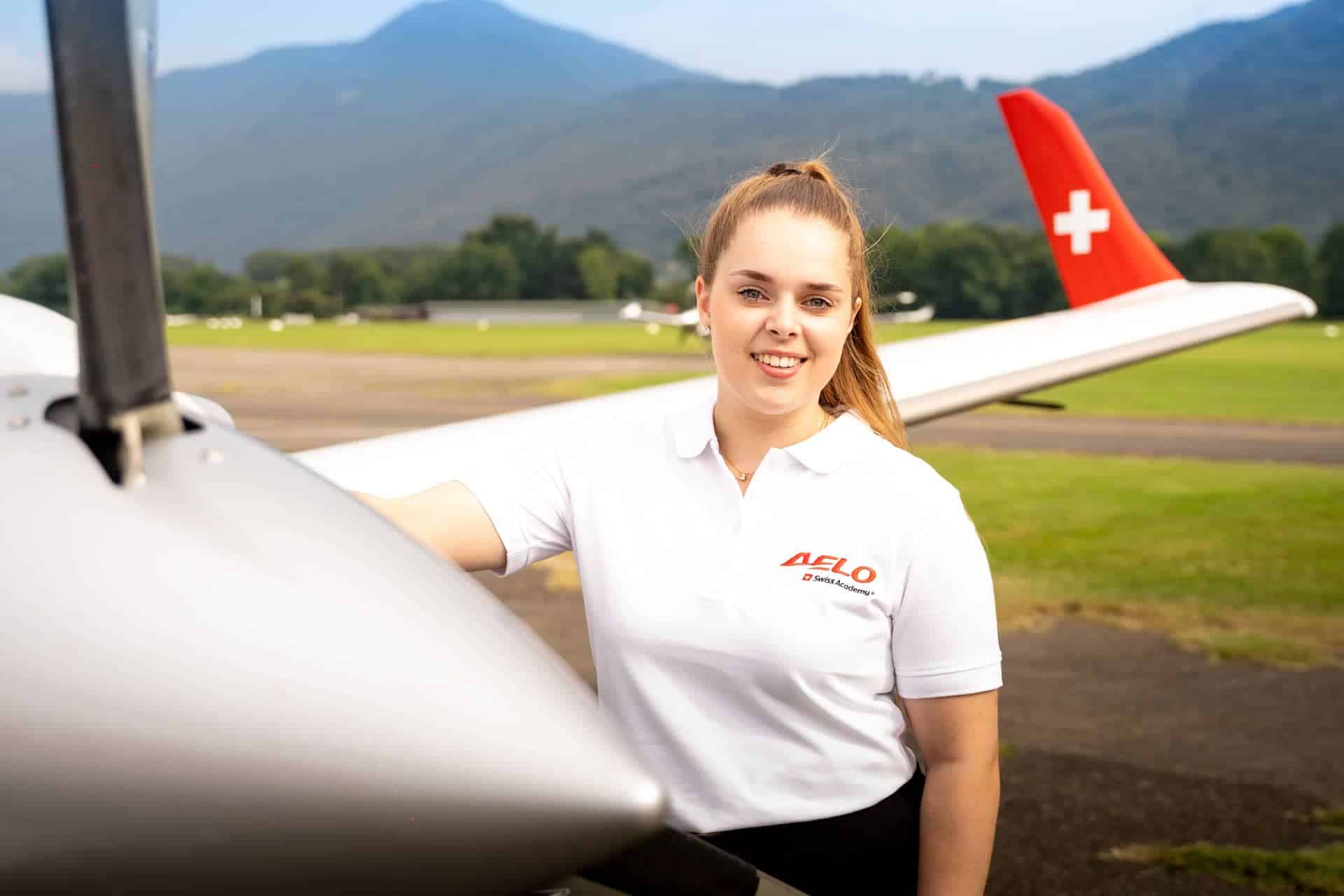Joven piloto sonriendo junto a avión AELO Swiss Academy con bandera suiza al fondo.